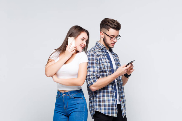 cute woman and handsome man in jeans shirts standing back to back and writing sms on smart phones isolated on white background - Fotografie, Obrázek