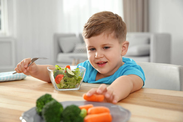 Adorable little boy eating vegetables and salad at table in room - Fotoğraf, Görsel