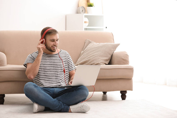 Young man with headphones and laptop sitting on floor in living room - Fotó, kép