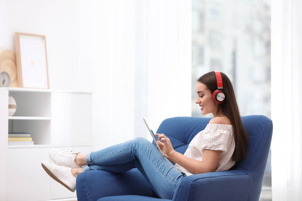 Young woman with headphones and tablet sitting in armchair at home - 写真・画像
