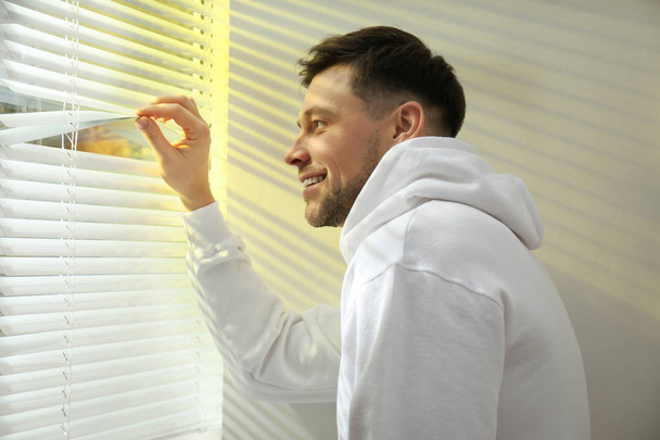 Handsome man opening window blinds at home - Фото, изображение