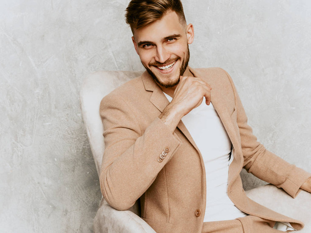 Portrait of handsome smiling hipster lumbersexual businessman model wearing casual beige suit. Fashion stylish man posing against gray wall sitting in chair in interior - Foto, afbeelding