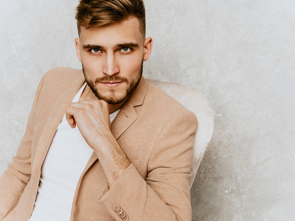 Portrait of handsome smiling hipster lumbersexual businessman model wearing casual beige suit. Fashion stylish man posing against gray wall sitting in chair in interior - 写真・画像
