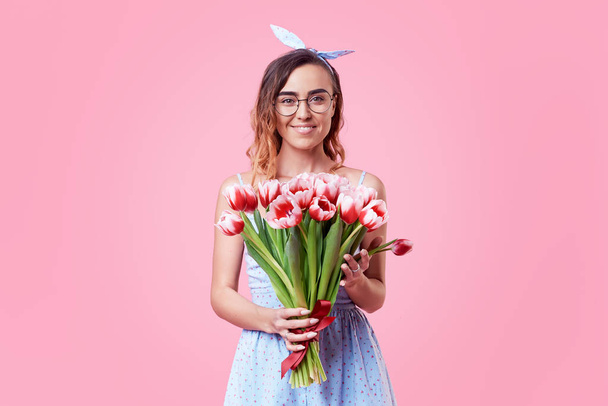 fröhliche junge Dame in Retro-runden Gläsern, die aufgeregt ist, am Frauentag vor rosa Hintergrund einen Strauß roter Frühlingsblumen zu bekommen. es schaut nach rechts, Raum für Text - Foto, Bild