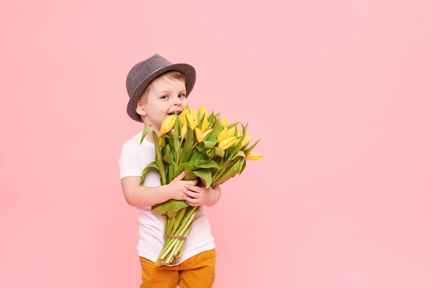 Adorable smiling child with spring flower bouquet looking at camera isolated on pink. Little toddler boy in hat holding yellow tulips as gift for mom - Foto, afbeelding