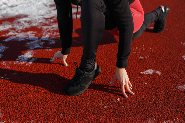 Detailní záběr dívka ve sportovním oblečení na trati pro běh na zasněženém stadionu se chystá běhat. běží od nízkého startu na zasněžený stadion. Fit a sportovní životní styl - Fotografie, Obrázek