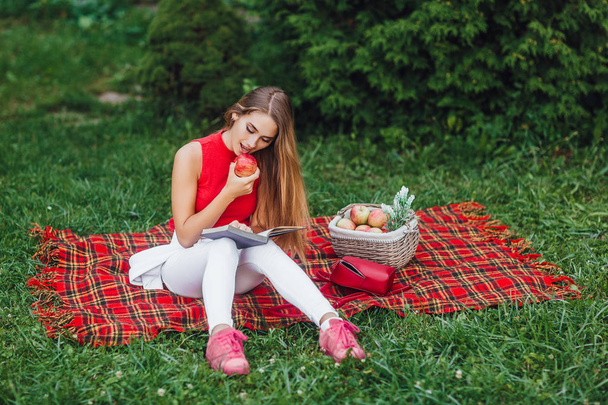 blonde woman reading book and eating apple in park - Фото, изображение