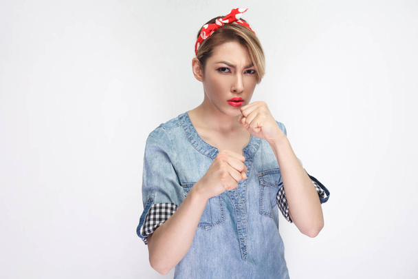 serious beautiful young woman in casual blue denim shirt with makeup and red headband standing with boxing fists and looking at camera isolated on white background. - Foto, Bild
