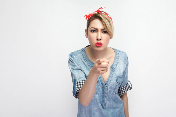 serious beautiful young woman in casual blue denim shirt with makeup and red headband standing and pointing at camera isolated on white background. - Φωτογραφία, εικόνα