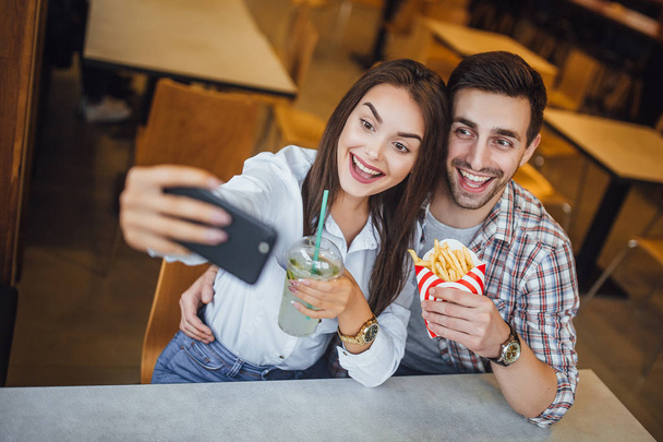 young couple making selfie on mobile phone with mojito and hamburger, smiling in fast food cafe, selective focus - Foto, Imagen