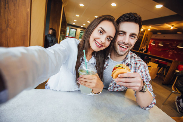 young couple making selfie on mobile phone with mojito and hamburger, smiling in fast food cafe, selective focus - Foto, imagen