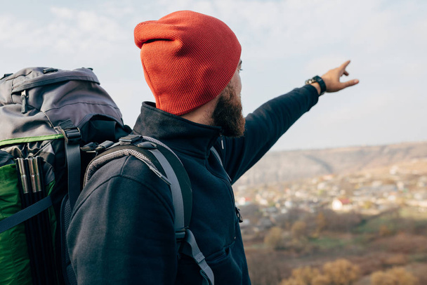 Imagen vista trasera de un joven excursionista con sombrero rojo, caminando por las montañas, mostrando algo en el horizonte. Viajero barbudo hombre relajante después del alpinismo. Viajes, personas y estilo de vida
 - Foto, imagen