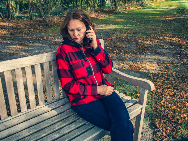 Σοβαρή μελαχρινή 50 χρονών, κάθεται σε ένα παγκάκι με ένα κινητό  - Φωτογραφία, εικόνα