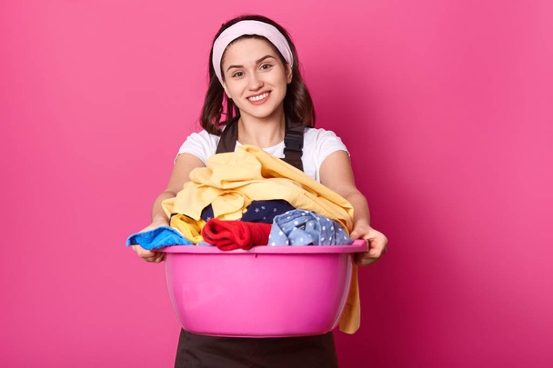 Young woman holds basin full of clean linen. Beautiful housewife looks happy after doing laundry. Smiling female works about house. Maid in brown apron enjoys fresh colourful linen on pink background. - Photo, Image