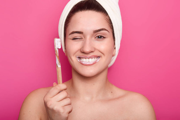 Beauty portrait of a smiling beautiful naked woman. Cheerful girl with towel on head holds toothbrush and winks. Young lady shows white teeth. Female with one closed eye isolated over pink background. - Фото, зображення
