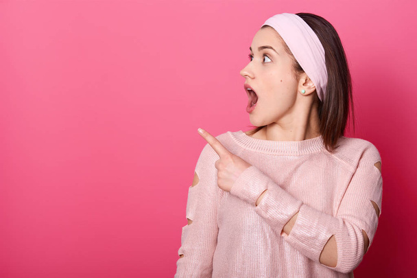 Portrait of shocked woman. Astonished girl stands isolated on pink studio background. Female wears rose clothes looks aside, points her fore finger to left upper corner. Facial expression concept. - Fotó, kép