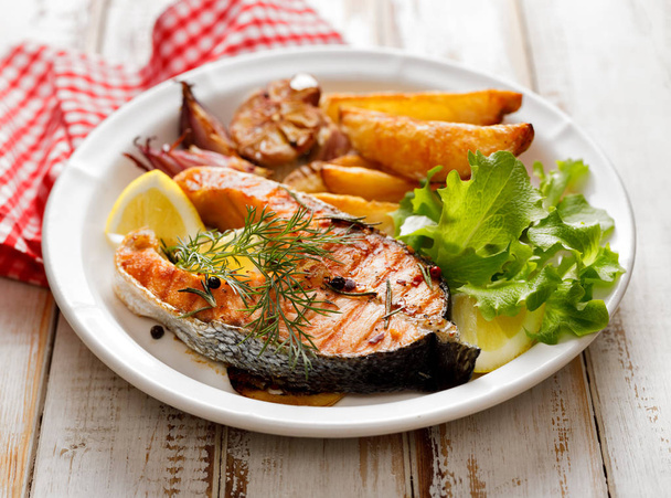 Стейк из лосося на гриле, часть лосося на гриле со свежим салатом и картофельными клинками на белой керамической тарелке на деревянном деревенском столе
 - Фото, изображение