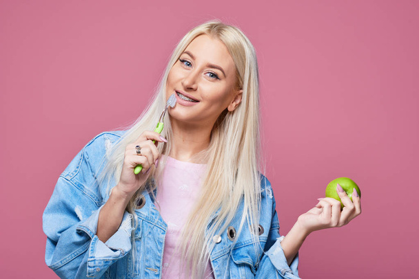 Close up portrait of young blond woman with braces on teeth holding green apple and toothbrush. The concept of oral hygiene after eating - Фото, изображение