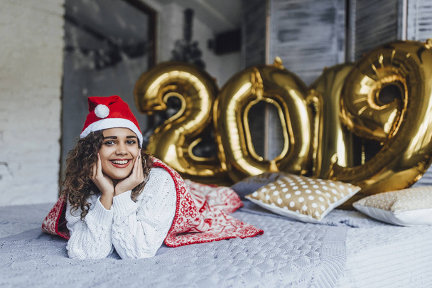 Young beautiful curly woman in Santa Clause hat wrapped in warm blanket with New Years gifts in loft room in background of 2019 numbers - Fotó, kép