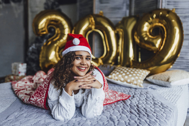 Young beautiful curly woman in Santa Clause hat wrapped in warm blanket with New Years gifts in loft room in background of 2019 numbers - Fotó, kép