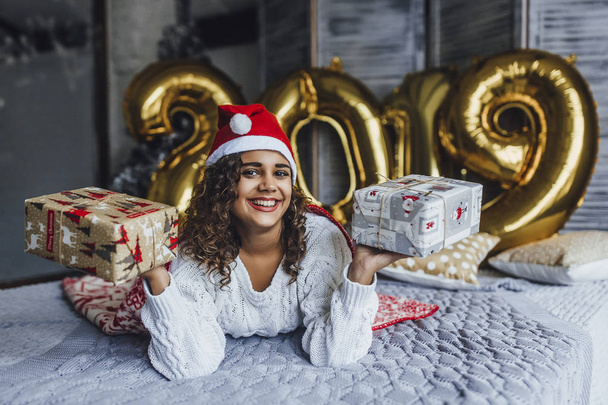Young beautiful curly woman in Santa Clause hat wrapped in warm blanket with New Years gifts in loft room in background of 2019 numbers - 写真・画像
