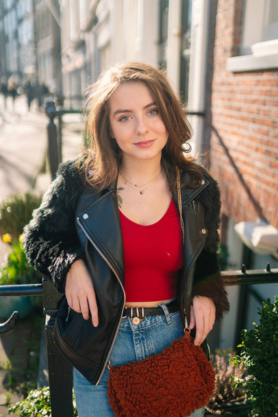 Portrait of a beautiful girl on a sunny day. Streets of Amsterdam. Great mood. A girl enjoys her lifestyle. She is wearing a red T-shirt, jeans and a leather jacket. - Foto, Imagem