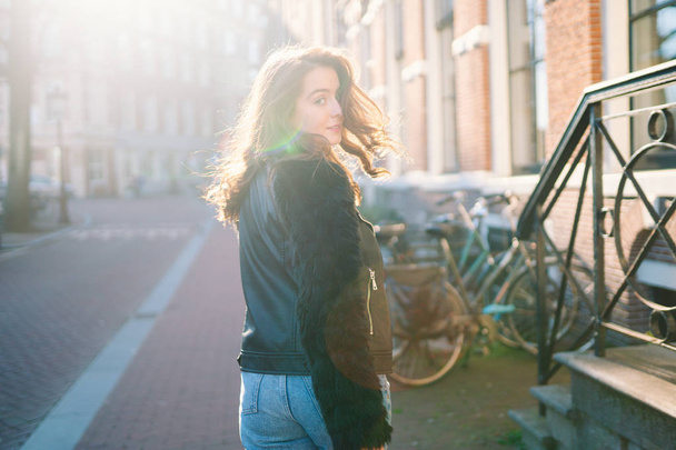 Portrait of a beautiful girl on a sunny day. Streets of Amsterdam. Great mood. A girl enjoys her lifestyle. She is wearing a red T-shirt, jeans and a leather jacket. - Foto, imagen