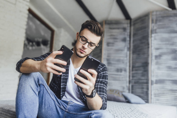 Young handsome blogger in glasses comparing two mobile phones in loft room - Photo, Image