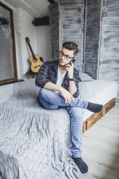 handsome man in glasses speaking on cellphone and posing at camera in loft style studio - Fotoğraf, Görsel
