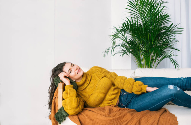 Happy young brunette in jeans and sweater dreaming on the sofa.  - Zdjęcie, obraz