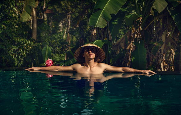 der Typ im Infinity-Pool. Pool im Dschungel. Urlaub auf Bali. der Kerl bräunt. Entspannung. konischer asiatischer Hut. - Foto, Bild