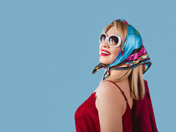 stylish smiling cheerful fancy blonde woman with make up in neckerchief and sunglasses on blue background - Photo, Image