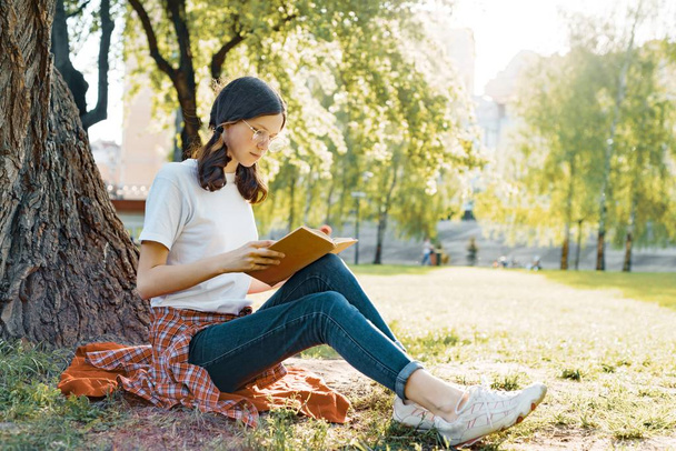Parkta çim üzerinde bir ağacın altında oturup kitap okumak gözlüklü kız öğrenci - Fotoğraf, Görsel