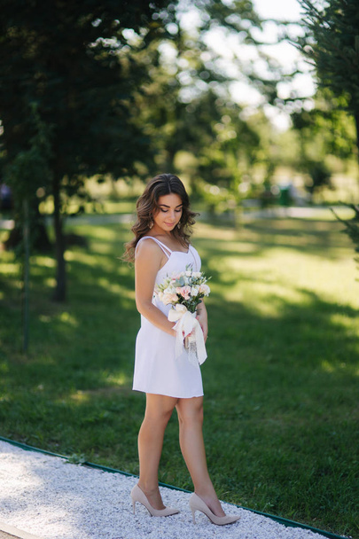 Elegant woman in beautiful white dress with bouquet of flowers walking in the park. Female model outside - Foto, imagen