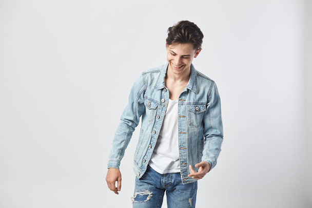 Smiling young dark-haired guy in a white t-shirt, jeans and a denim jacket stands on the white background  in the studio - Photo, image