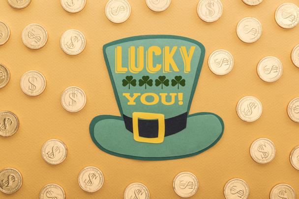 top view of golden coins with dollar signs near green paper hat with lucky you lettering on orange background  - Valokuva, kuva