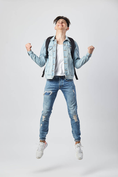 Young dark-haired guy with black backpack  on his shoulder dressed in a white t-shirt, jeans and a denim jacket happily jumps on the white background in the studio - Foto, Imagem