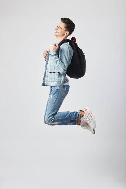 Young dark-haired guy with black backpack  on his shoulder dressed in a white t-shirt, jeans and a denim jacket happily jumps on the white background in the studio - Photo, image