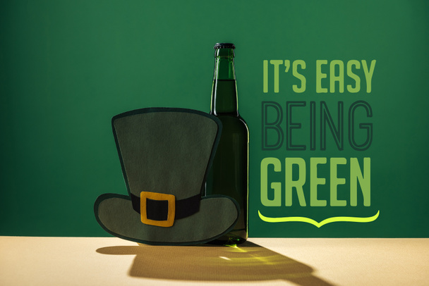 Bierflasche und Papierhut in der Nähe seines leicht grünen Schriftzugs auf grünem Hintergrund - Foto, Bild