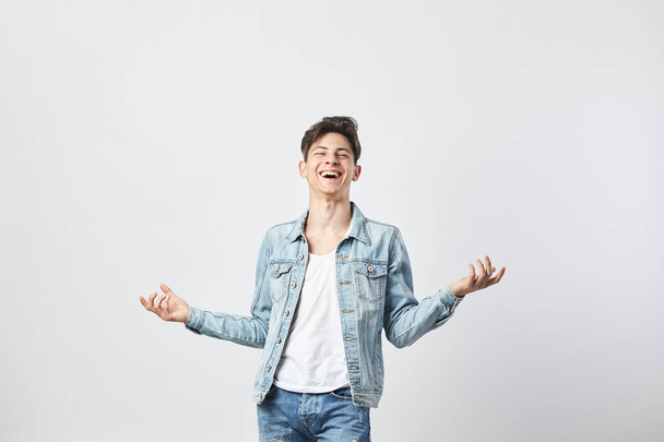 Laughing dark-haired guy dressed in a white t-shirt, jeans and a denim jacket spreads his arms to the side on the white background in the studio - Fotografie, Obrázek