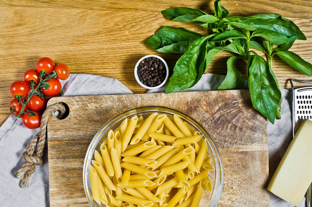 Pasta Penne, ingredients for cooking. Cherry tomatoes, Basil, Parmesan, pepper. Wooden background, top view - Zdjęcie, obraz