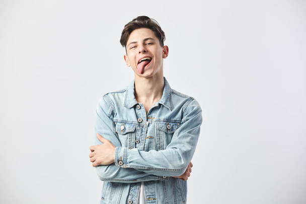 Funny dark-haired guy dressed in a white t-shirt and a denim jacket shows his tongue on the white background in the studio - Φωτογραφία, εικόνα