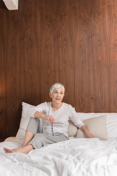 Cheerful senior woman with big beautiful smile sitting on comfy bed  - Fotó, kép