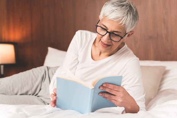 mature woman with glasses lying on comfy bed and reading book  - Φωτογραφία, εικόνα