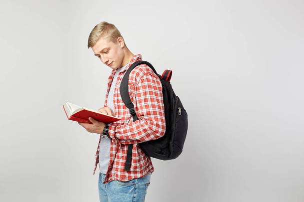 Young blond guy with black backpack on his shoulder dressed in a white t-shirt, red checkered shirt and jeans reads a book on the white background  in the studio - Fotó, kép