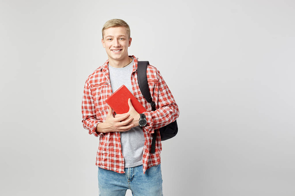 Smiing young blond guy with black backpack on his shoulder dressed in a white t-shirt, red checkered shirt and jeans holds books  in his hands on the white background  in the studio - Foto, imagen