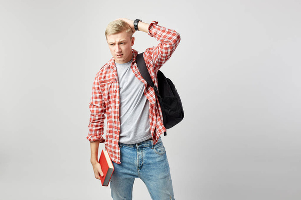 Blond guy with black backpack on his shoulder dressed in a white t-shirt, red checkered shirt and jeans holds book in his hand and other hand on his head on the white background in the studio - Valokuva, kuva