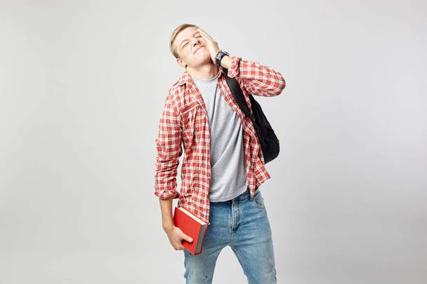 Blond guy with black backpack on his shoulder dressed in a white t-shirt, red checkered shirt and jeans holds book in his hand and other hand on his face on the white background in the studio - Foto, imagen