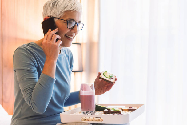 Joyful senior woman talking on mobile phone and eating healthy breakfast on bright sunny morning in her apartment - Foto, imagen