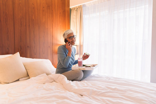 Radosny starszy kobieta rozmowy na telefon komórkowy i jedzenie zdrowe śniadanie na jasny słoneczny poranek w jej mieszkaniu - Zdjęcie, obraz